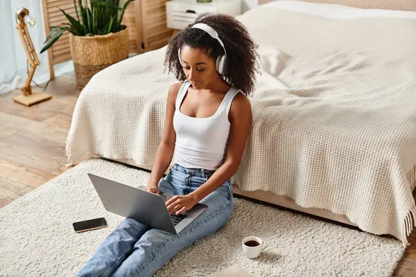 A curly African American woman in a tank top sits on the floor using a laptop in a modern bedroom. — Stock Photo