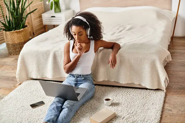A curly African American woman in a tank top sits on the floor using a laptop in a modern bedroom setting. — Stock Photo