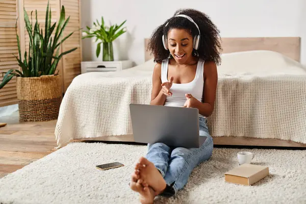 Una mujer afroamericana en una camiseta sin mangas se sienta en el suelo, centrada en su computadora portátil en un entorno de dormitorio moderno. — Stock Photo