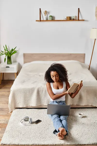 Mujer afroamericana rizada en camiseta sin mangas sentada en el suelo, profunda en la lectura de un libro en un entorno de dormitorio moderno. - foto de stock