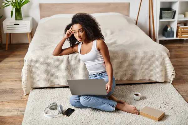 A curly African American woman in a tank top sits on the floor, engrossed in using a laptop in a modern bedroom. — Stock Photo