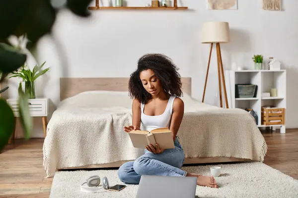 Eine lockige Afroamerikanerin in einem Tank-Top sitzt auf dem Boden und liest in einem modernen Schlafzimmer ein Buch. — Stockfoto