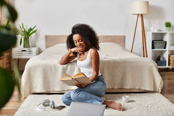 Riccio donna afroamericana in canotta seduta sul pavimento assorbita nella lettura di un libro accattivante in una camera da letto moderna. — Foto stock