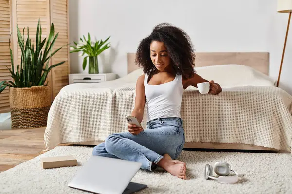 Mujer afroamericana rizada sentada en el suelo junto a la computadora portátil en el dormitorio moderno. - foto de stock