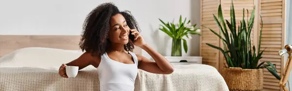 Curly African American woman in tank top chatting on cell phone while seated on modern bed. — Stock Photo