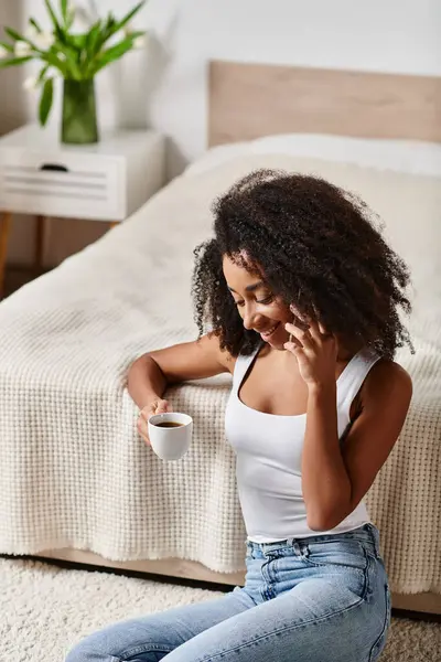 Mujer afroamericana rizada en una camiseta sin mangas sentada en el suelo, absorta en una llamada telefónica en un dormitorio moderno. - foto de stock