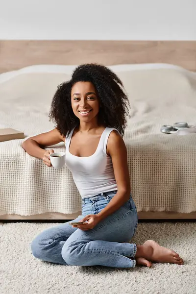 Curly African American woman in a tank top sitting on the floor in front of a bed in a modern bedroom. — Stock Photo