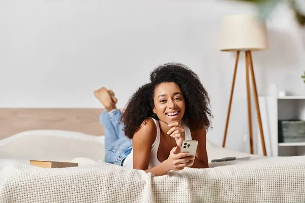 A curly African American woman in a tank top lays on a bed, holding a cell phone in a modern bedroom. — Stock Photo