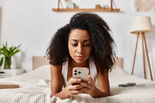 Mujer afroamericana rizada en una camiseta sin mangas, acostada en una cama, absorta en su teléfono celular en un dormitorio moderno. - foto de stock