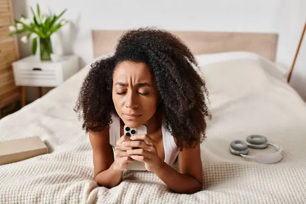 Mulher afro-americana encaracolada em um tampo de tanque, deitada em uma cama segurando um telefone celular em um quarto moderno. — Fotografia de Stock