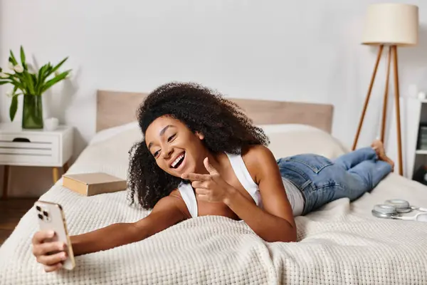 Curly African American woman in tank top reclining on bed, engrossed in cell phone. — Stock Photo