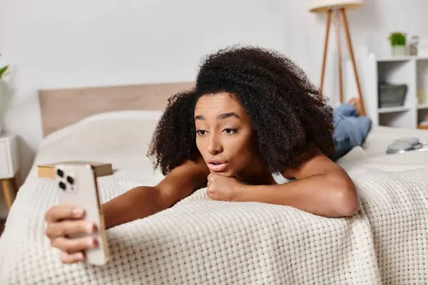 Curly African American woman in tank top laying on bed, holding cell phone in modern bedroom. — Stock Photo