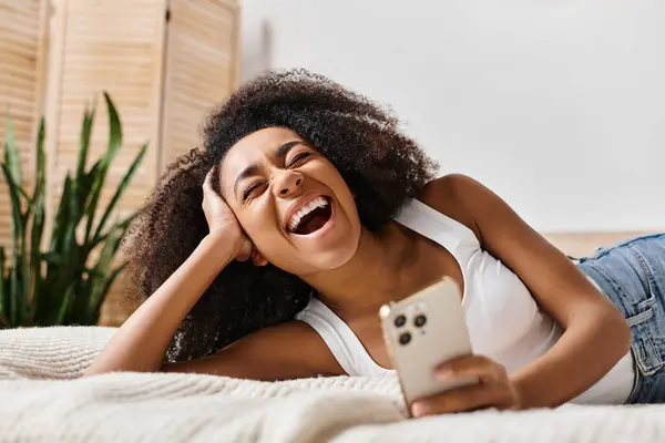 Curly African American woman in a tank top relaxing on a bed, holding a smartphone in a modern bedroom. — Stock Photo