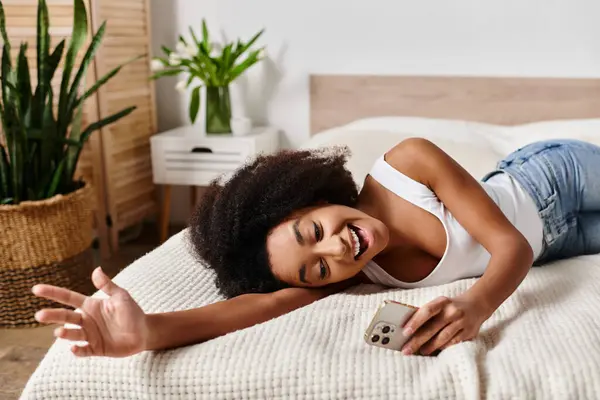 A curly African American woman in a tank top relaxes on a bed next to a vibrant green plant in a modern bedroom. — Stock Photo