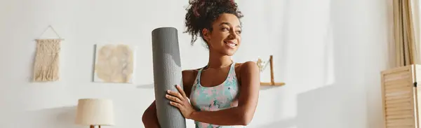 Curly African American woman in active wear holding a yoga mat in a serene room, ready for a relaxing workout. — Stockfoto