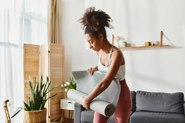 Donna afroamericana riccia in leggings rosa con un tappetino da yoga, pronta per la sua routine di allenamento a casa. — Foto stock