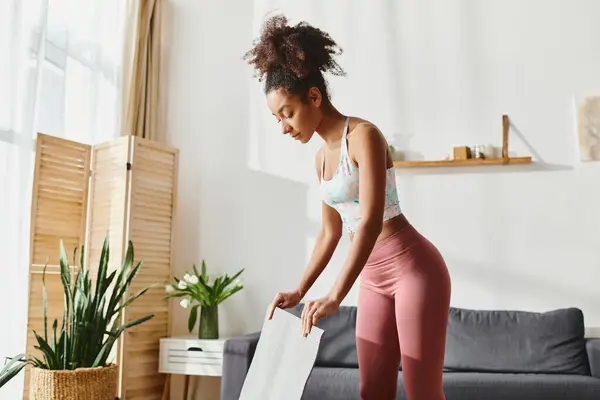 Una donna afroamericana riccia in leggings rosa con un tappetino da yoga mentre si allena a casa. — Foto stock