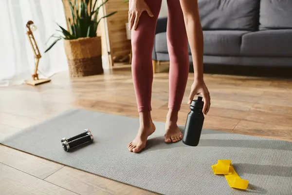 Cropped African American woman in activewear standing on yoga mat, holding a bottle of water. - foto de stock
