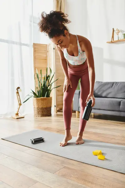 Lockige Afroamerikanerin in Aktivkleidung steht auf einer Yogamatte und hält eine Flasche Wasser zur Flüssigkeitszufuhr in der Hand. — Stockfoto