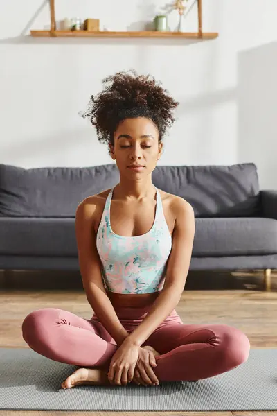 Curly African American woman in activewear peacefully sitting on a yoga mat in a cozy living room, practicing mindfulness. — Stock Photo