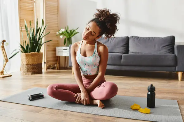 Femme afro-américaine frisée en tenue active s'assoit sur un tapis de yoga, pratiquant la pleine conscience dans un cadre confortable salon. — Photo de stock