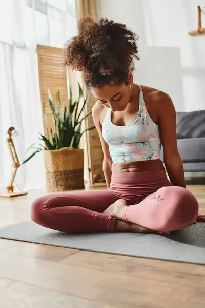Кудрявая афроамериканка в активной одежде сидит на коврике для йоги в уютной гостиной, сосредоточившись на внимательности и расслаблении. — стоковое фото