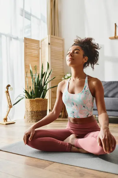 Mujer afroamericana rizada en ropa activa sentada en una esterilla de yoga en una acogedora sala de estar, practicando ejercicio consciente. - foto de stock