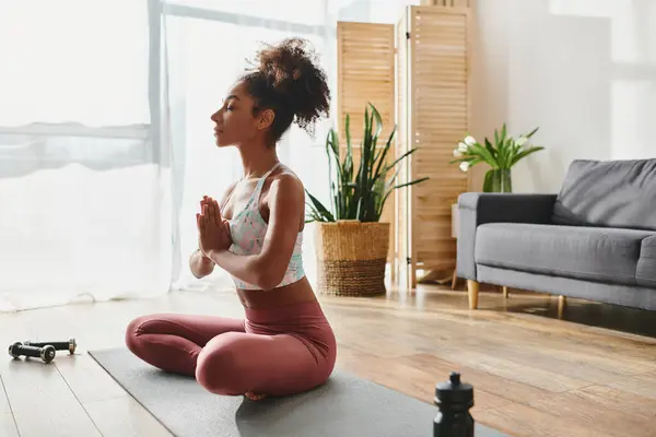 Une afro-américaine frisée en vêtements de sport assise sur un tapis de yoga dans un salon confortable, axée sur sa pratique du yoga. — Photo de stock