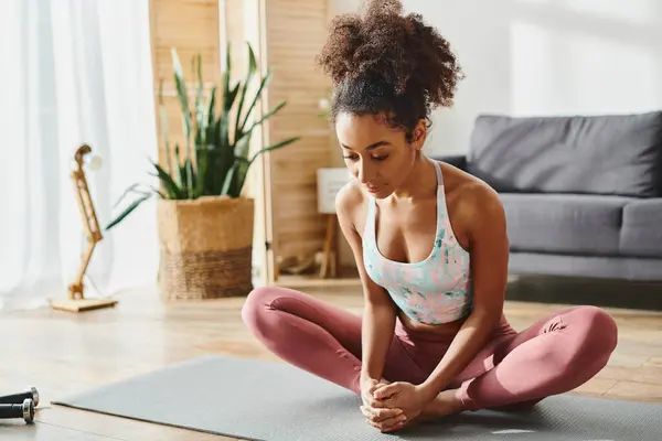 Curly mulher afro-americana em desgaste ativo sentado em um tapete de ioga em uma sala de estar aconchegante, encontrar a paz através da prática de ioga. — Fotografia de Stock