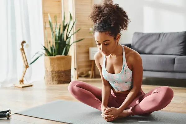 Curly African American woman in active wear sitting on a yoga mat in a cozy living room, finding peace through yoga practice. — Stock Photo