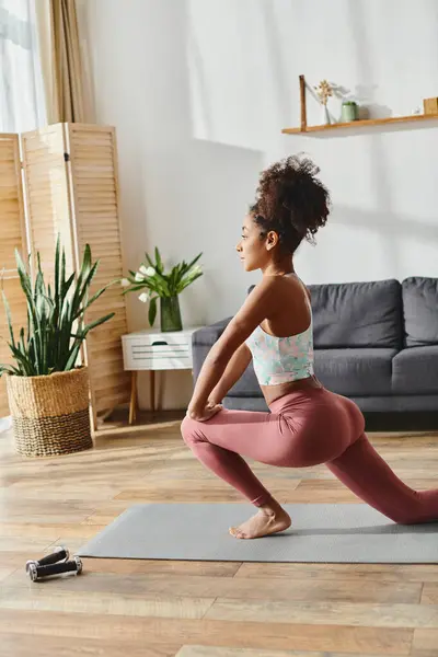 Une femme afro-américaine frisée en vêtements de sport pratique le yoga dans son salon confortable rempli de lumière naturelle. — Photo de stock