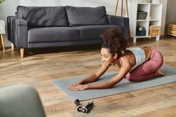 Lockige Afroamerikanerin in aktiver Kleidung hält zu Hause eine Yoga-Pose auf einer Yogamatte. — Stockfoto