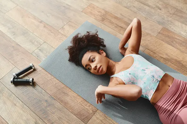 Curly African American woman in activewear laying on yoga mat for home workout. - foto de stock