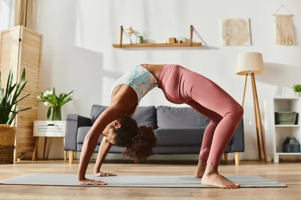 Кудрявая афроамериканка в активной одежде изящно выполняет стойку на руках на коврике для йоги в спокойной домашней обстановке. — стоковое фото