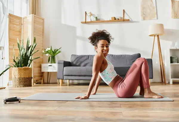 Eine Afroamerikanerin mit lockigem Haar, in Activwear gekleidet, führt anmutig eine Yoga-Pose auf einer Yogamatte zu Hause aus. — Stockfoto