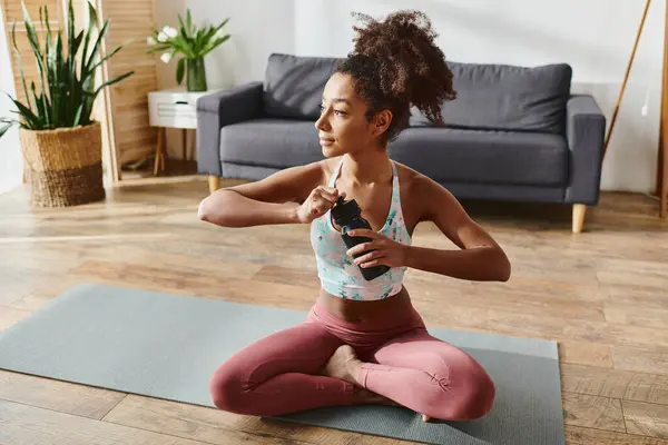 Riccio afroamericano donna in attivo usura prende una pausa per idratarsi con una bottiglia d'acqua mentre seduto su un tappetino yoga. — Foto stock