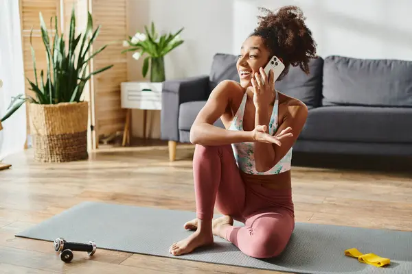Una donna afroamericana riccia in abito attivo si siede su un tappetino yoga, parlando su un telefono cellulare a casa. — Foto stock
