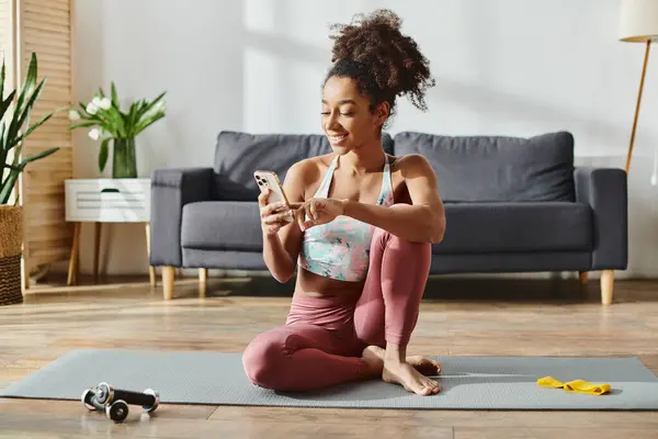 Curly African American woman in activewear sits on yoga mat, engrossed in phone screen. — Stock Photo