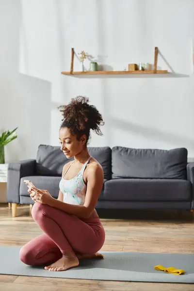 Une afro-américaine frisée en tenue active s'assoit sur un tapis de yoga, absorbée dans son téléphone. — Photo de stock