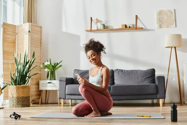 A curly African American woman in activewear sitting on a yoga mat, engrossed in her phone screen while working out at home. — Stock Photo