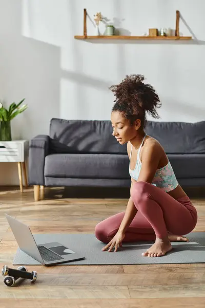 La donna afroamericana riccia in abbigliamento attivo si siede sul tappetino yoga con il computer portatile davanti, impegnandosi in una sessione di fitness online. — Foto stock