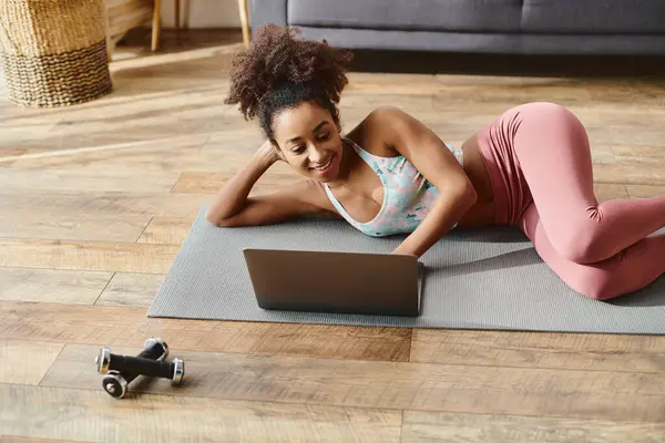 Curly African American woman in activewear doing yoga on a mat, using a laptop. — Stock Photo