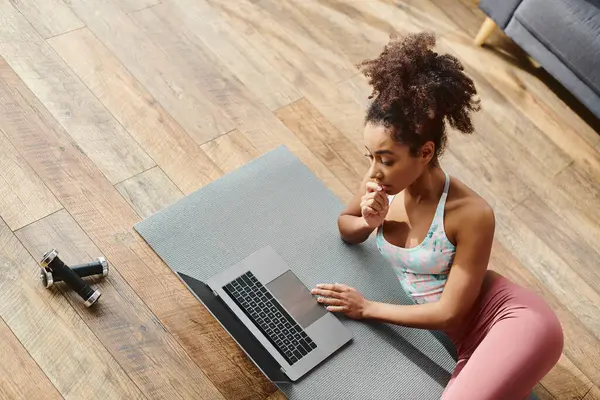 Active curly African American woman practices yoga while typing on laptop on a yoga mat at home. — Stock Photo