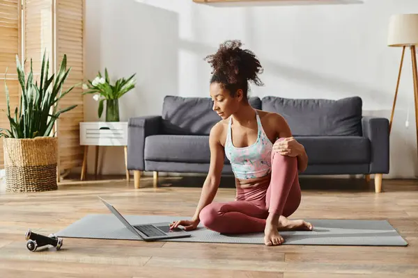 A curly African American woman in active wear peacefully sitting on a yoga mat in a cozy living room. — Stock Photo