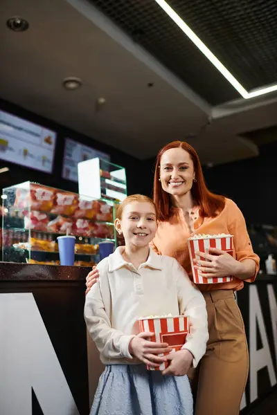 A woman stands next to a daughter, holding two boxes of popcorn, while enjoying a family trip to the cinema. — Stock Photo