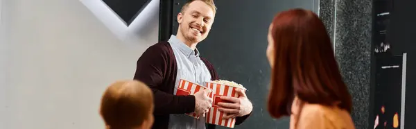 Man joyfully holds a box of popcorn in front of a woman, both smiling happily, at a cinema with their family. — Stock Photo