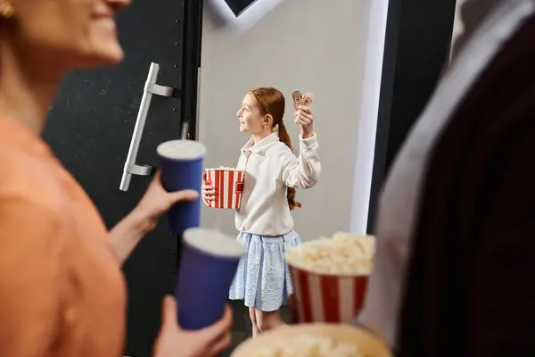 Ребенок счастливо держит чашку попкорна, стоя рядом с семьей в кинотеатре. — стоковое фото