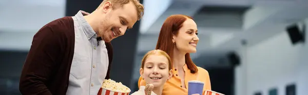 Мужчина стоит рядом с женой и ребенком, оба улыбаются, а она держит коробку попкорна в кинотеатре. — стоковое фото