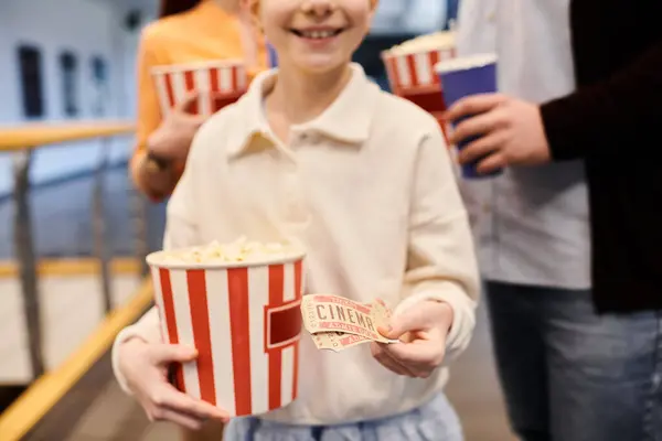 Girl joyfully holds bucket of popcorn and tickets in a cinema, surrounded by his happy family. — Stock Photo