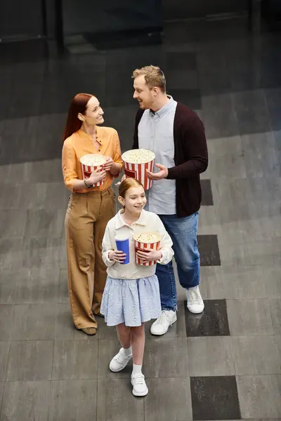 Family happily hold popcorn buckets while standing next to a little girl, enjoying a movie night at the cinema. — Stock Photo